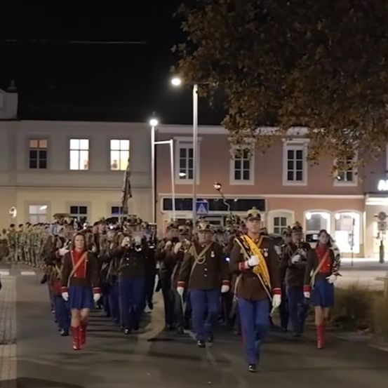 Ein Militärorchester marschiert am Abend, gekleidet in Uniformen und mit Instrumenten, vor einem Gebäude mit mehreren Fenstern.