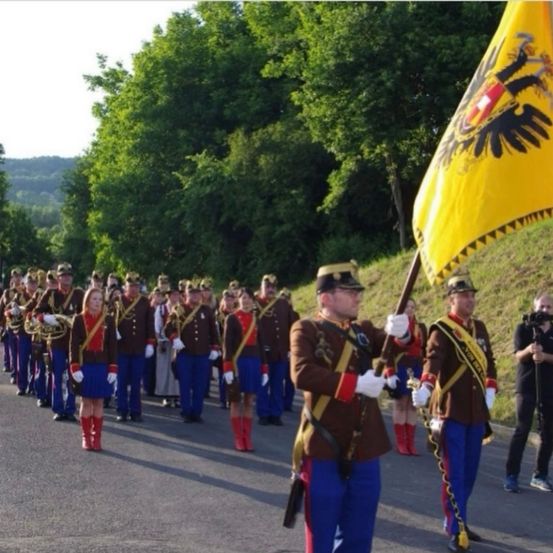 Eine Musikkapelle zieht auf der Straße. Sie tragen Uniformen und rote Stiefel. Der Mann vorne trägt eine Fahne. Hinter ihnen filmt ein Mann die Parade mit einer Kamera.