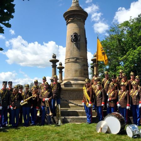 Eine Band in Uniformen steht vor einem Denkmal mit einer gelben Fahne. Sie halten Musikinstrumente, einschließlich Trompeten und Trommeln. Das Denkmal hat Statuen und Verzierungen darauf.
