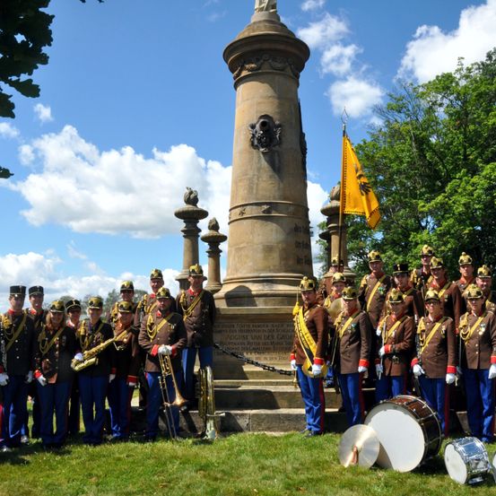 Eine Band in Uniformen steht vor einem Denkmal mit einer gelben Fahne. Sie halten Musikinstrumente, einschließlich Trompeten und Trommeln. Das Denkmal hat Statuen und Verzierungen darauf.