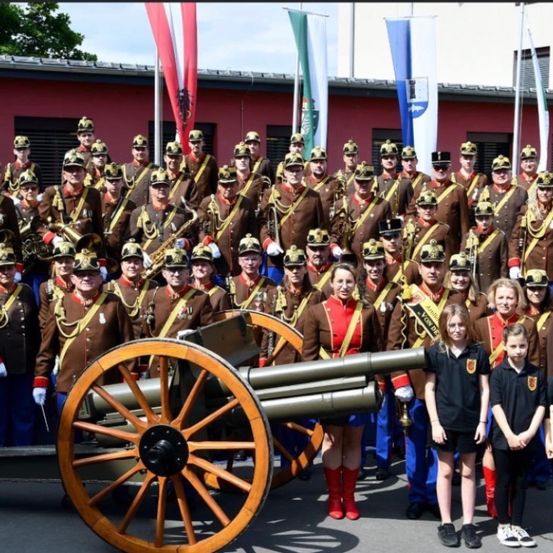Eine Marschkapelle in Uniform posiert mit einer Kanone, mit Kindern in der Nähe, vor dem Hintergrund von Flaggen und einem roten Gebäude.