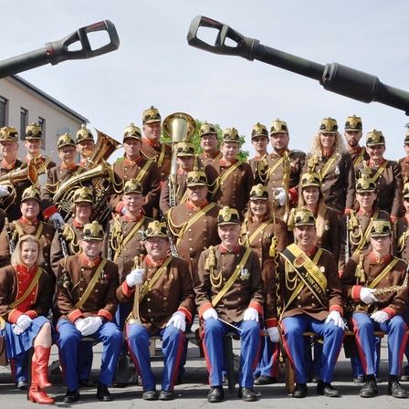 Eine Militärkapelle in braunen Uniformen mit weißen Handschuhen und Mützen posiert vor zwei großen Kanonen. Männer und Frauen sitzen und stehen mit verschiedenen Instrumenten.