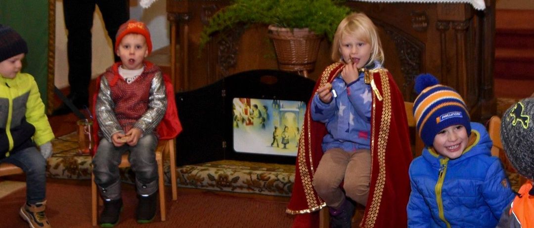 Ein Mann in einer schwarzen Jacke steht bei einem Podest mit einem grünen Banner. Kinder in Kostümen sitzen auf Stühlen. Ein Tisch mit Kerzen und Pflanzen steht dahinter.