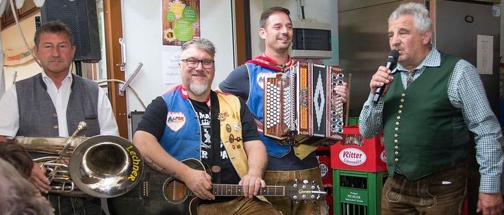 Vier Männer spielen in einem Raum, einer hält ein Mikrofon, ein anderer spielt Akkordeon und ein Dritter spielt Gitarre. Ein Poster an der Wand mit der Aufschrift '23.08.2014'.