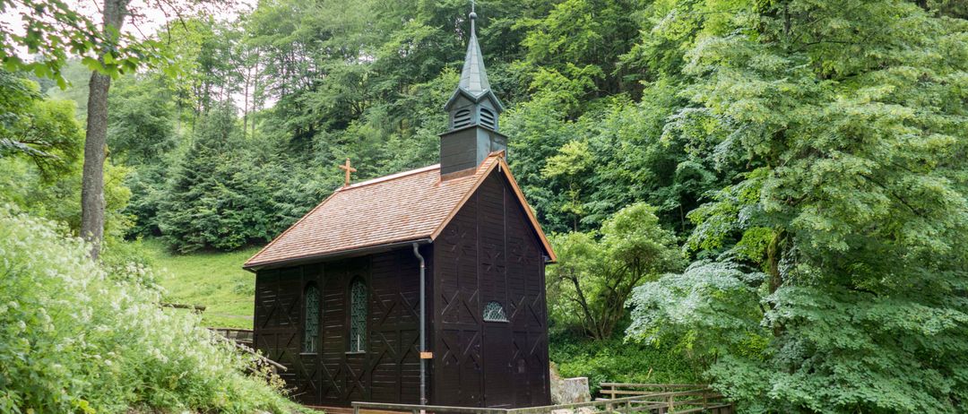 Eine kleine Holzkapelle mit einem spitzen Turm und einem braunen Dach, eingebettet in einem dichten grünen Wald. Die Kapelle ist von einem Holzzaun umgeben, mit einem Holzweg, der zu ihr hinaufführt.