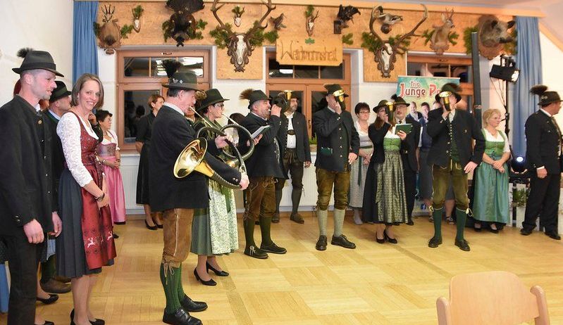 Men und Frauen in traditioneller bayerischer Kleidung versammeln sich in einem Raum mit Holzboden und Hirschköpfen an der Wand, einige blasen Hörner und andere schauen in ein Buch.