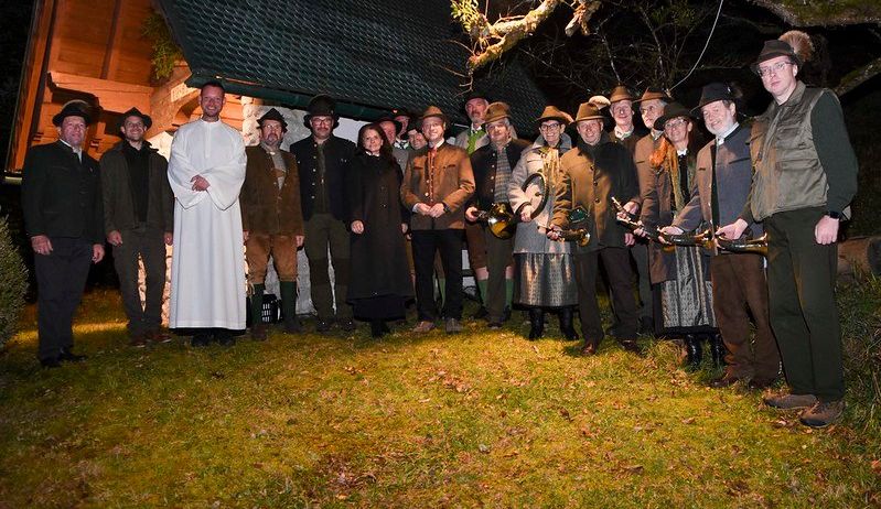 Eine Gruppe von Personen, einige in traditioneller Kleidung, versammelt sich bei Nacht vor einem Gebäude und posiert für ein Foto. Ein Priester steht unter ihnen, und sie halten Musikinstrumente.