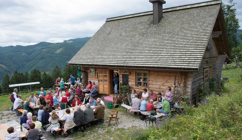 Eine Gruppe von Menschen sitzt um Tische herum vor einem rustikalen Blockhaus mit Schornstein. Einige stehen in der Nähe des Eingangs. Die Umgebung ist eine grasbewachsene Fläche mit einem Bergrücken im Hintergrund.