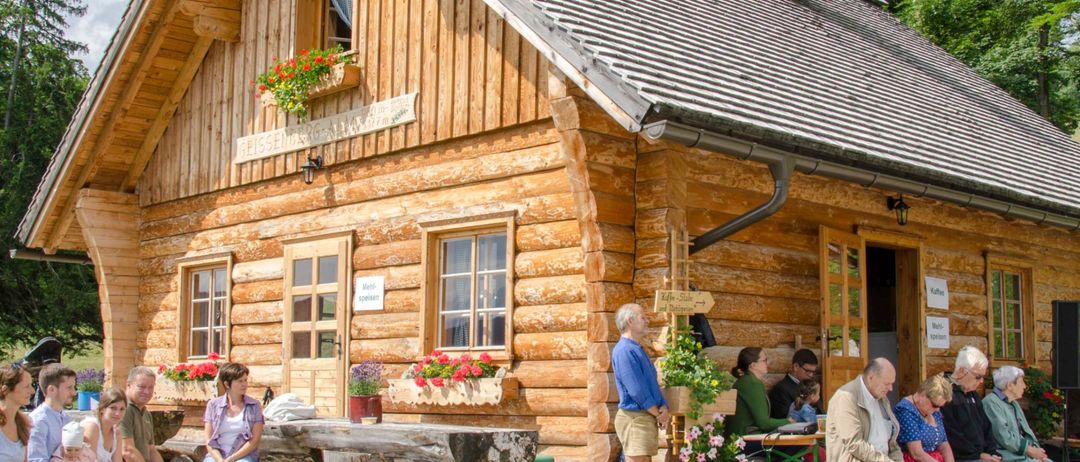 Eine Gruppe von Menschen sitzt um ein Holzhaus mit Blumen draußen. Ein Mann steht in der Nähe der Tür, und ein Tisch mit Blumen ist vor dem Haus platziert.