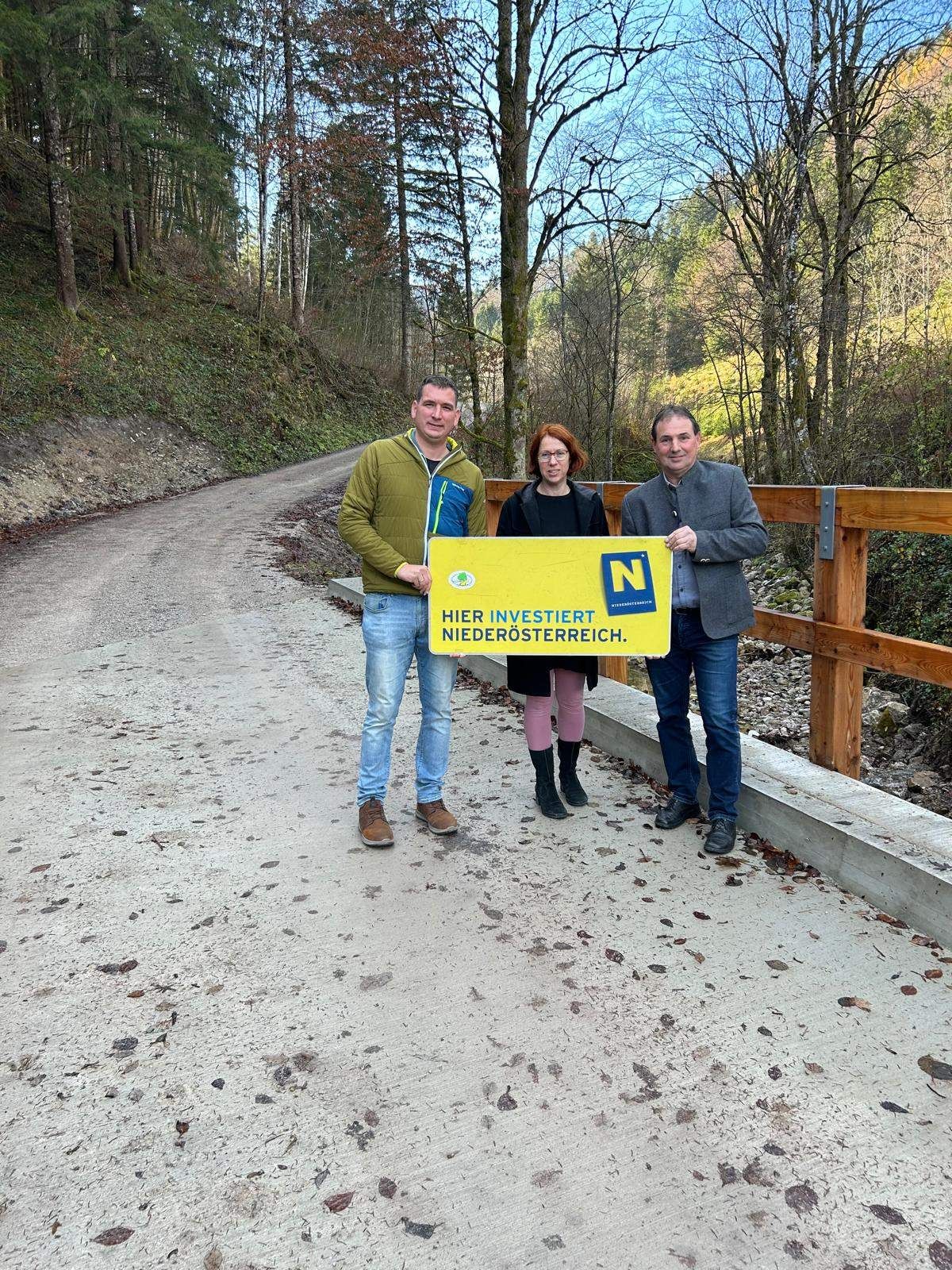 Drei Personen stehen vor einem gelben Schild, lächelnd, mit einer Holzbrücke und einer unbefestigten Straße dahinter.