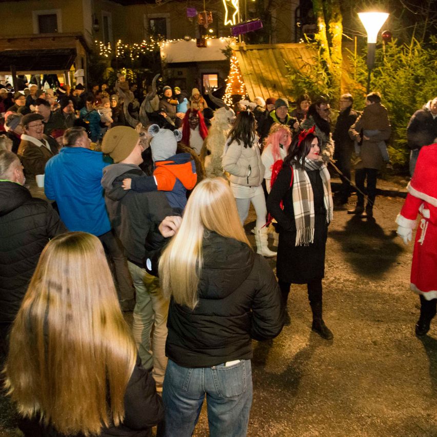 Eine Menschenmenge versammelt sich bei Nacht im Freien. Einige tragen Winterkleidung, Mützen und Schals. Eine Frau in einem Weihnachtsmannkostüm hält eine Leine. Eine Frau mit rotem Haar und einem karierten Schal spricht die Menge an. Eine Lampe und Gebäude mit Weihnachtsdekoration sind im Hintergrund.