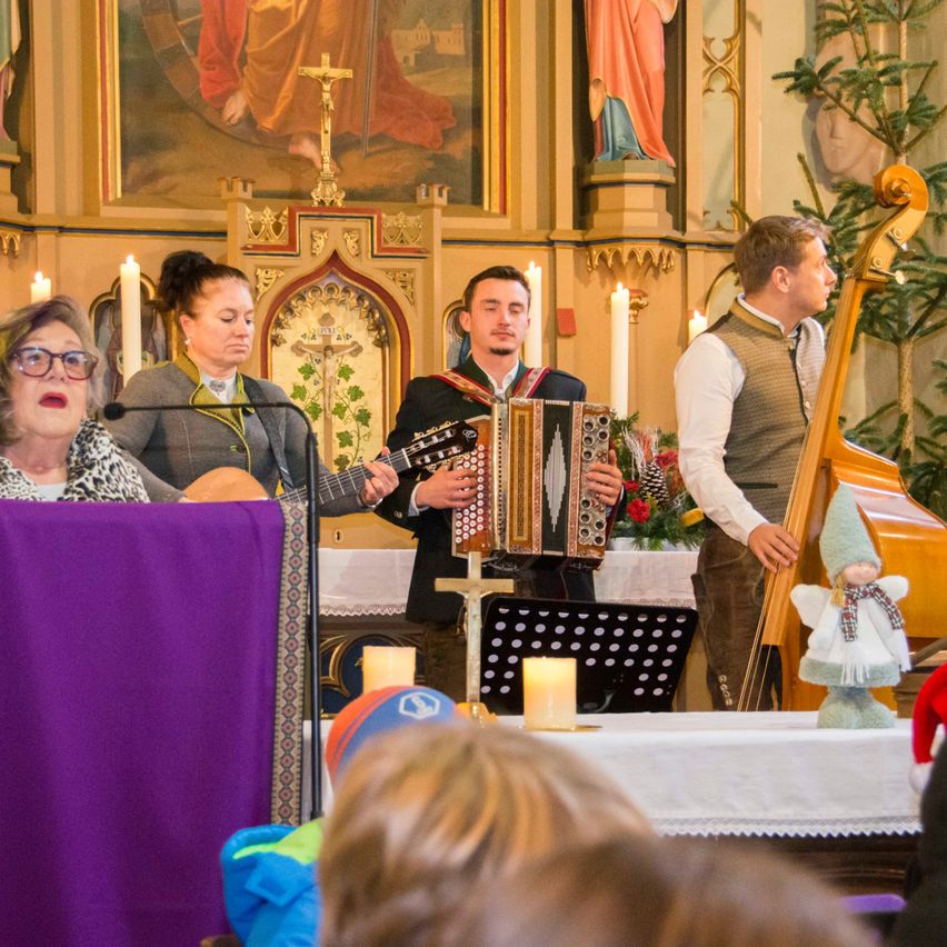 Eine Gruppe von Musikern tritt in einer Kirche mit traditionellen Instrumenten auf. Zwei Männer spielen Akkordeon und Cello, während eine Frau die Gitarre spielt. Kerzen und ein Weihnachtsbaum sind im Hintergrund zu sehen.