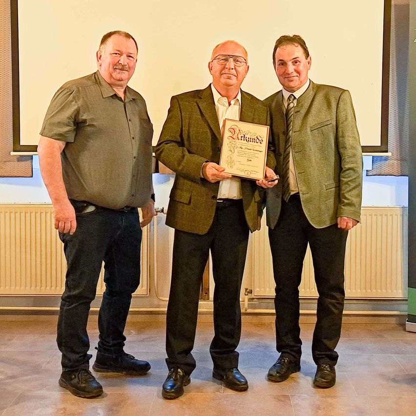 Drei Männer stehen vor einem weißen Bildschirm. Der Mann in der Mitte hält ein gerahmtes Zertifikat. Der Mann auf der linken Seite trägt ein grünes Hemd, während der Mann auf der rechten Seite eine grüne Jacke trägt.