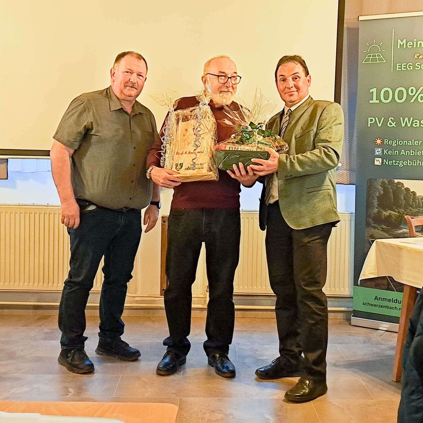 Drei Männer stehen zusammen drinnen, zwei von ihnen halten ein Geschenk in Plastikfolie. Der Mann in der Mitte trägt eine Brille und eine Krawatte. Ein Banner im Hintergrund steht für '100% PV & Was'.
