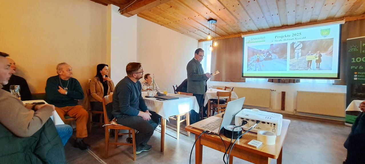 Ein Mann präsentiert ein Papier vor einer Projektionsleinwand mit einem Bild einer Baustelle. Mehrere Personen sitzen an Tischen.