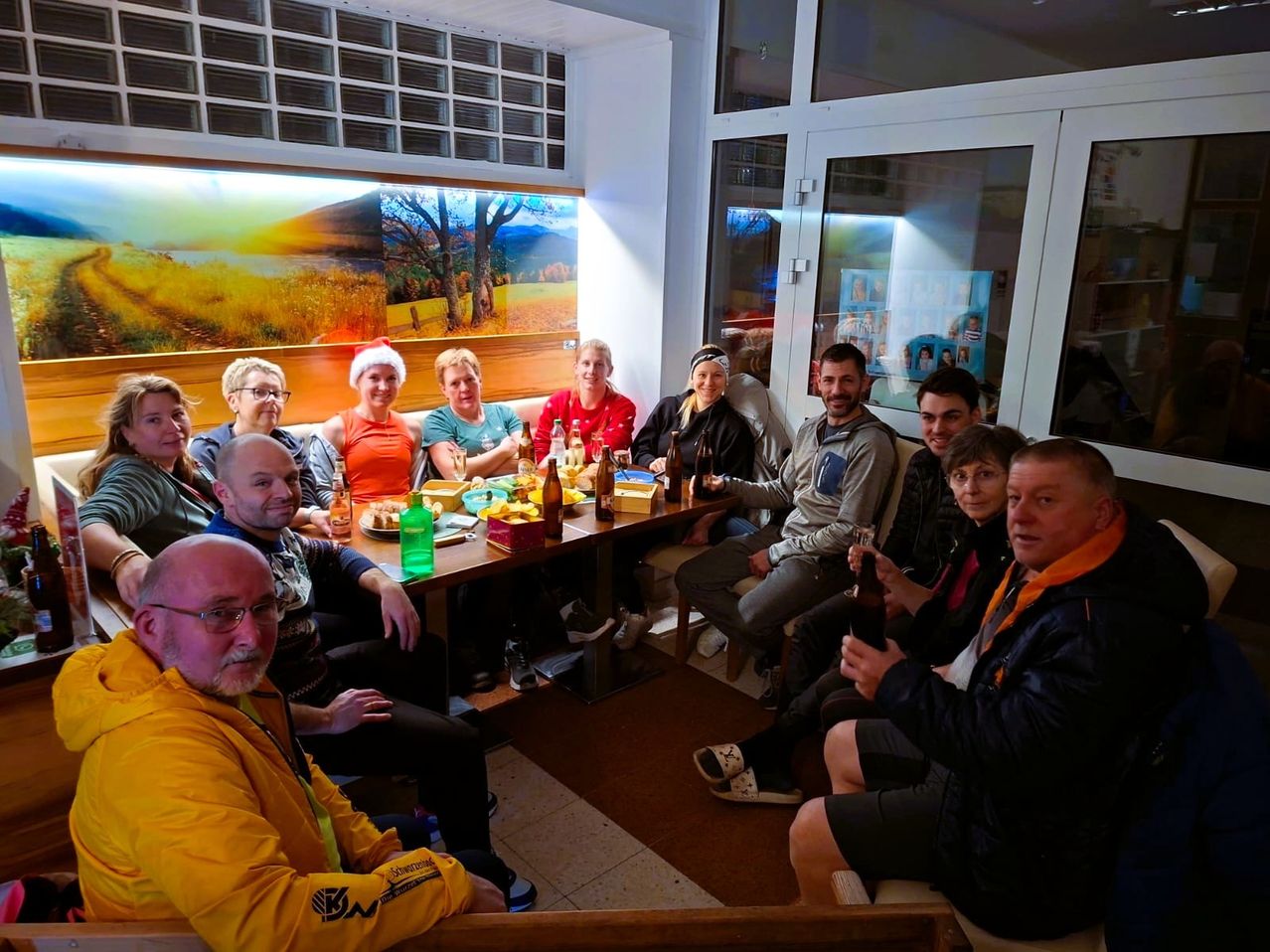 Eine Gruppe von Menschen sitzt in einem Esszimmer mit einem Tisch voller Essen und Getränke. Eine Frau mit einer Weihnachtsmütze sitzt am Tisch. Ein Gemälde einer Landschaft mit Bäumen und Bergen hängt an der Wand.