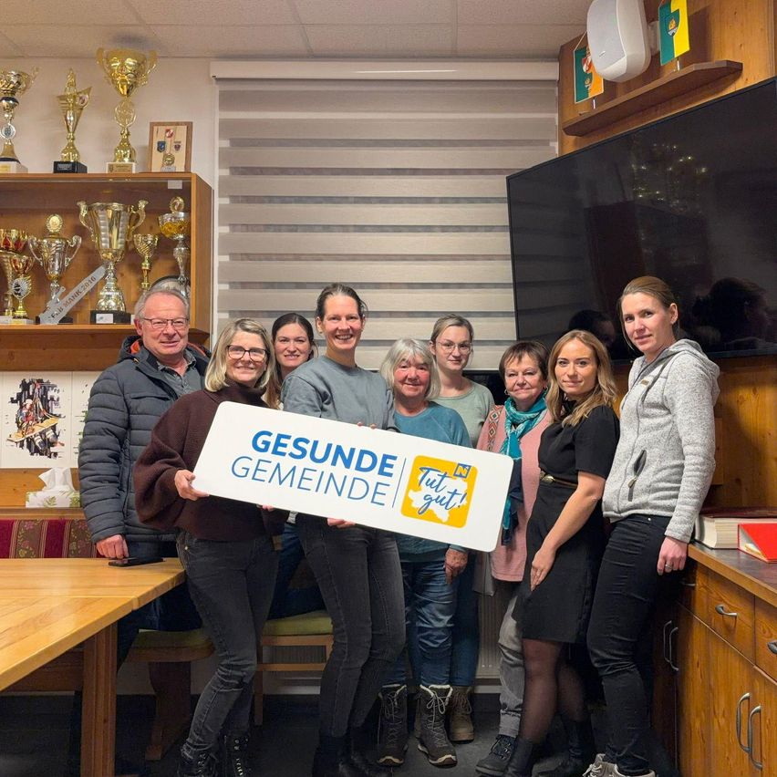 Eine Gruppe von Menschen steht in einem Raum und hält ein weißes Schild mit der Aufschrift 'Gesunde Gemeinde' und einem Logo. Sie tragen Freizeitkleidung, und einige tragen Brillen. Hinter ihnen befinden sich Regale mit Trophäen, ein Fernseher und ein Lautsprecher. Eine Frau in einer Kapuzenjacke hält ein Buch.