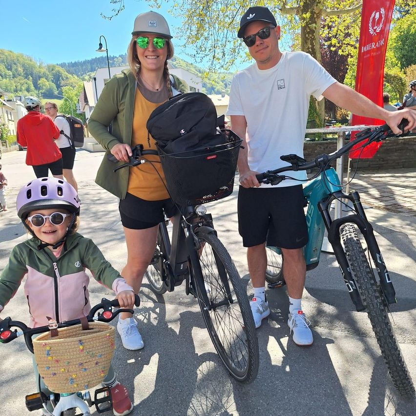 Eine Familie steht mit ihren Fahrrädern in einem Park. Eine Frau hält ein Kind auf einem Fahrrad. Ein Mann steht neben ihnen. Sie alle lächeln. Dahinter sind Menschen zu Fuß und Gebäude sichtbar.