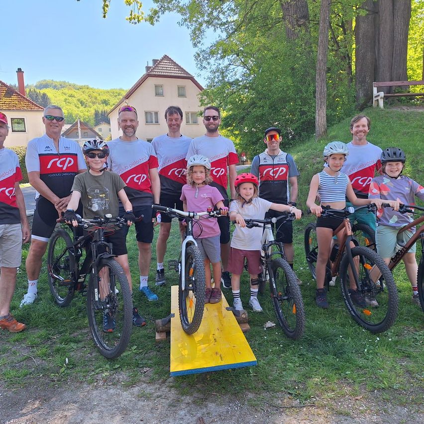 Eine Gruppe von Menschen posiert mit ihren Fahrrädern auf einem Rasen, trägt Helme und Fahrradtrikots. Bäume und Häuser im Hintergrund.