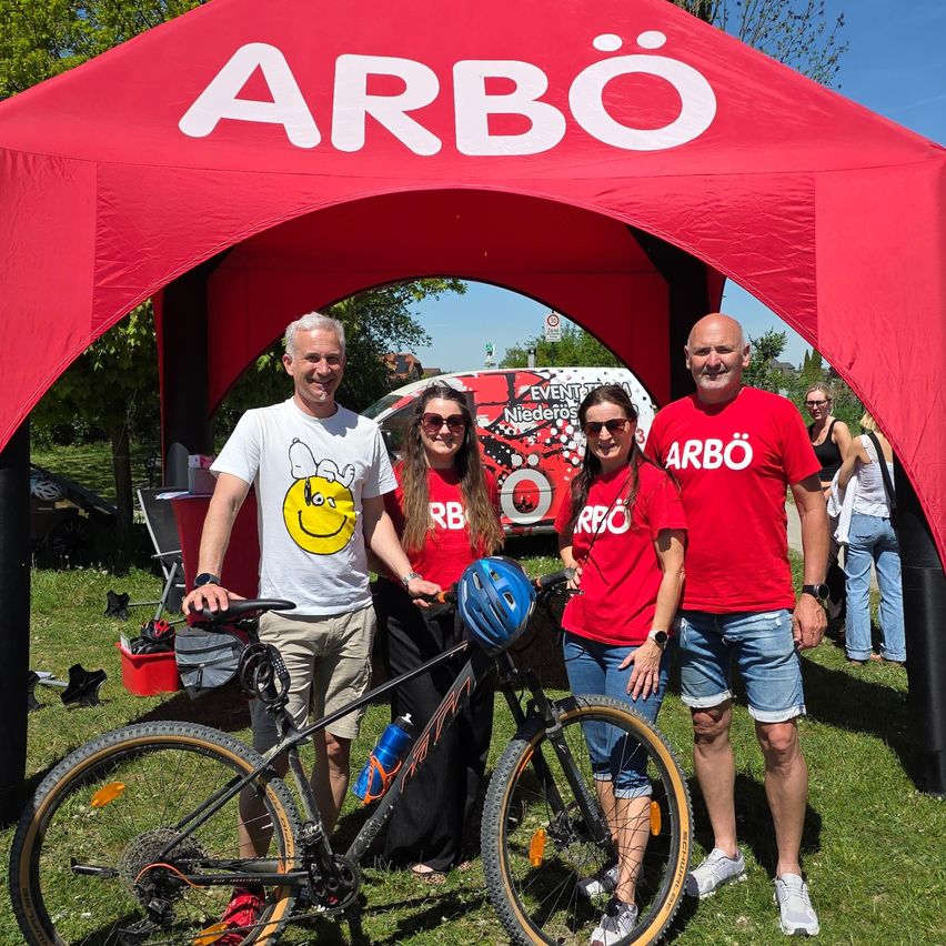 Vier Personen posieren mit einem Fahrrad unter einem roten ARBO-Zelt. Sie tragen rote T-Shirts, eine Frau hält einen Helm. Im Hintergrund ist ein Van geparkt, und Bäume und Menschen sind sichtbar.