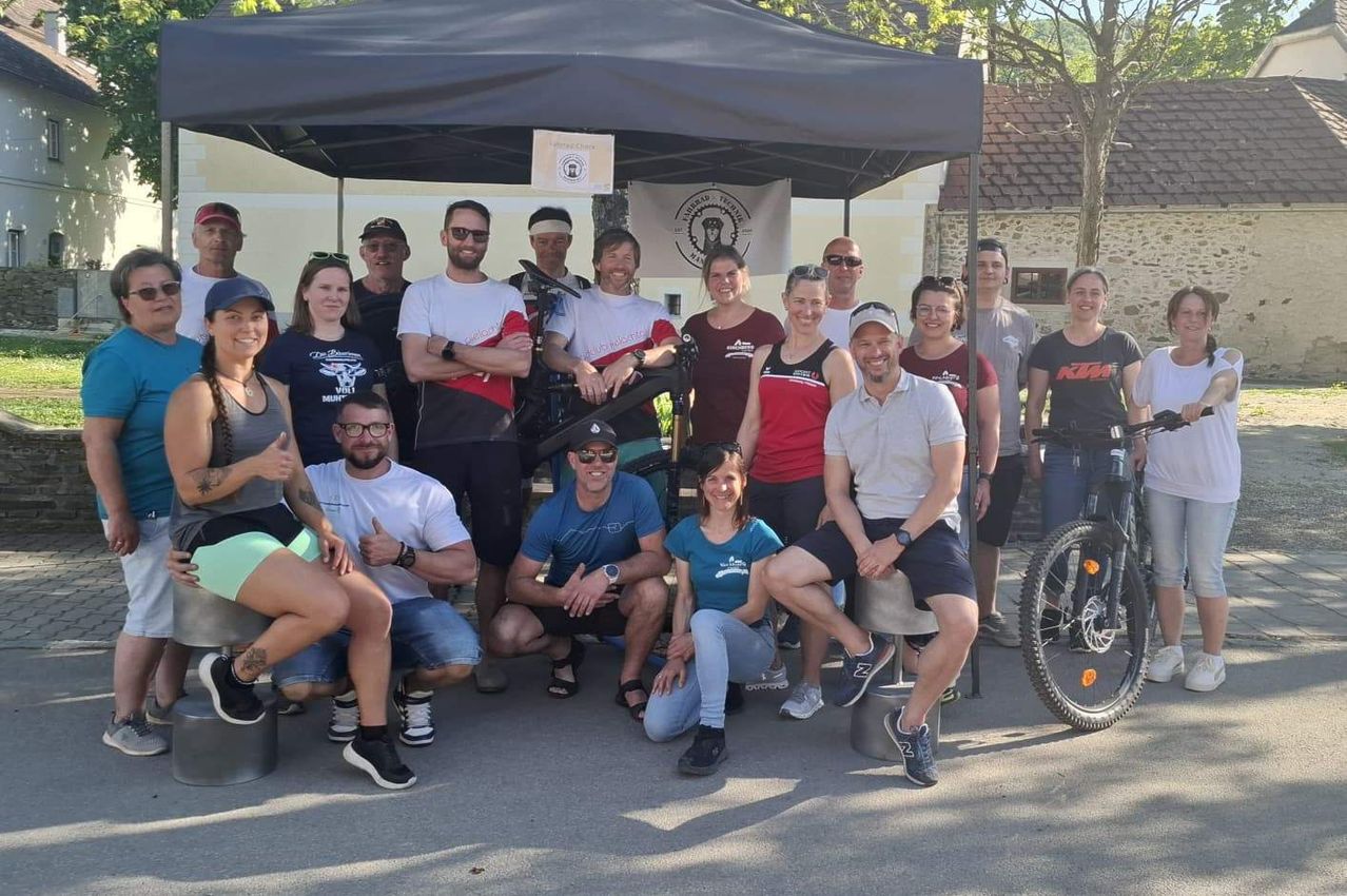 Eine Gruppe von Menschen in unterschiedlicher Kleidung und mit Accessoires posiert für ein Foto unter einem Zelt mit einem Banner. Einige halten Fahrräder.