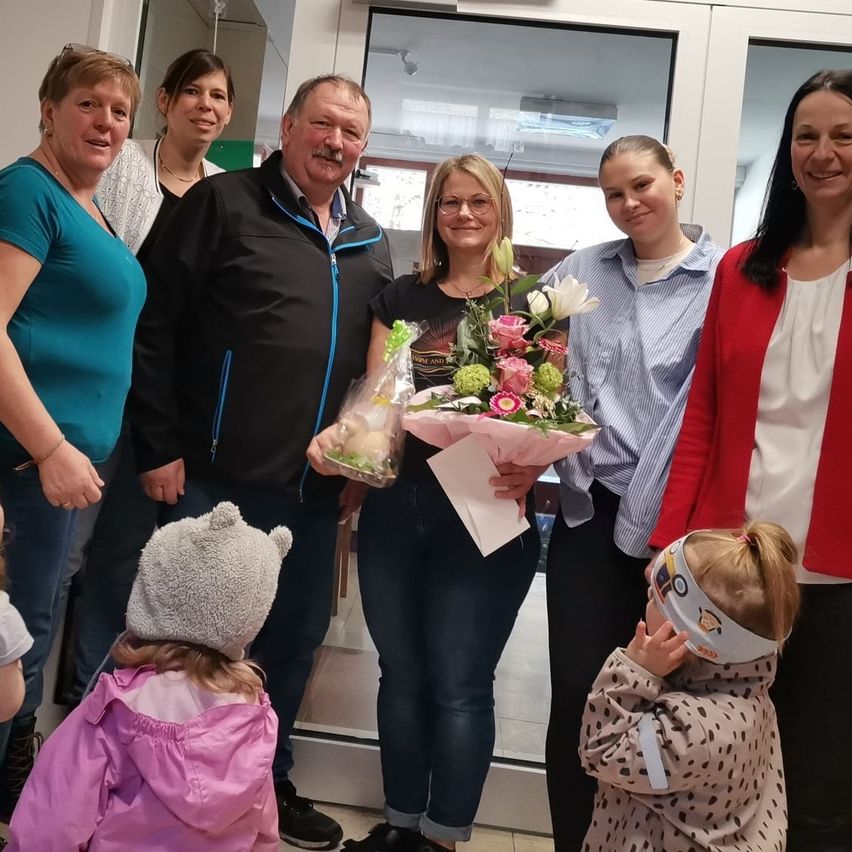 Eine Gruppe von Menschen steht lächelnd drinnen zusammen. Eine Frau hält einen Blumenstrauß und ein Mann ein Geschenk. Zwei junge Mädchen sind anwesend, eines mit einer Gesichtsmaske.