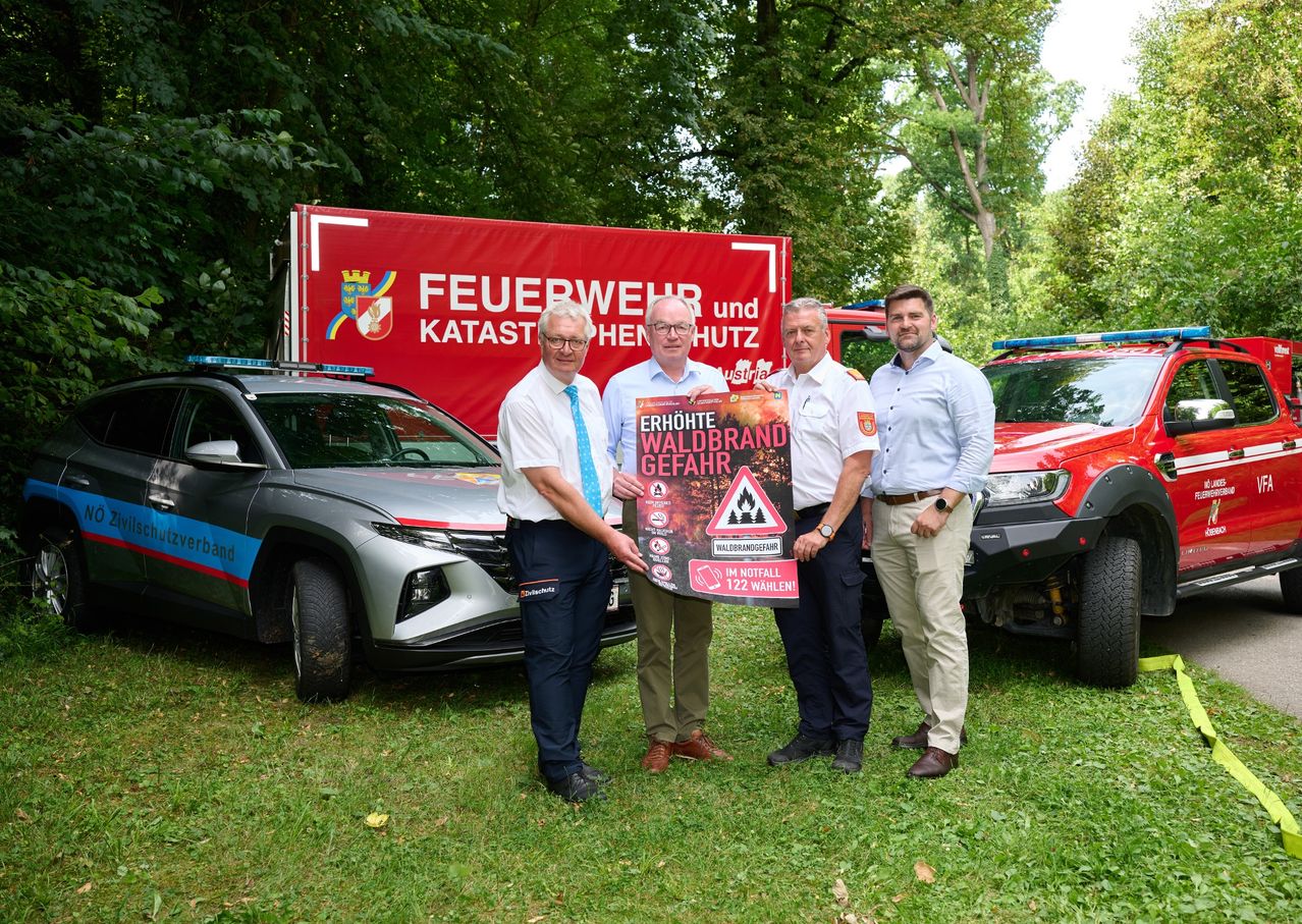 Vier Männer stehen vor einem Banner und zwei Feuerwehrautos. Der Mann in der Mitte hält ein Schild mit einer Warnung vor Waldbrandgefahr.