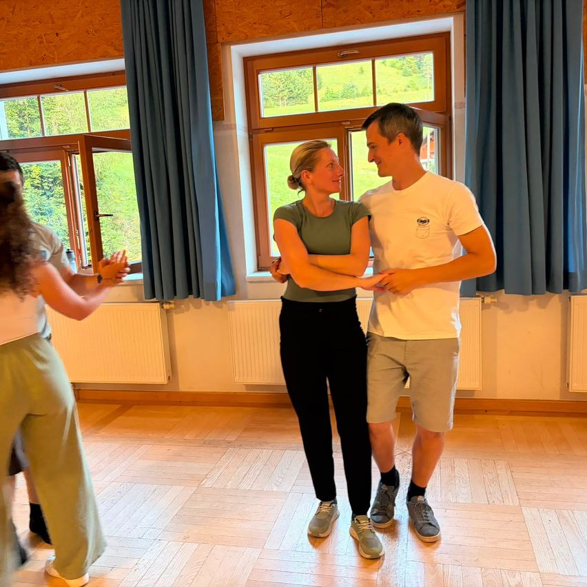 Ein Mann und eine Frau tanzen in einem Raum mit Holzboden und großen Fenstern, während ein anderes Paar im Hintergrund tanzt.