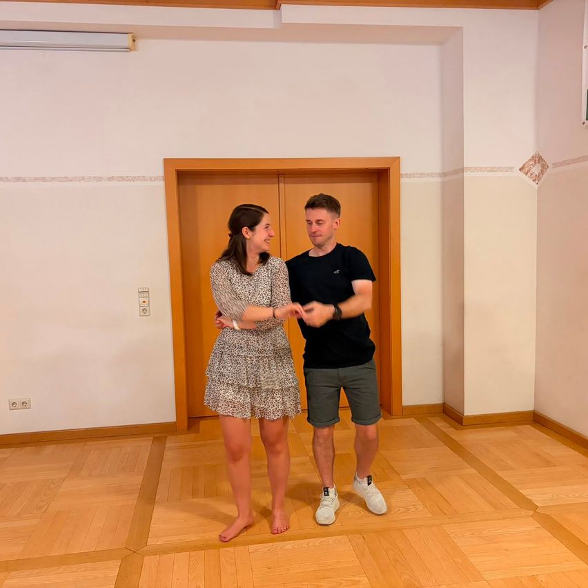 Ein Mann und eine Frau stehen in einem Raum, lächeln und halten Händchen. Sie sind beide barfuß. Der Raum hat Holzböden und weiße Wände.