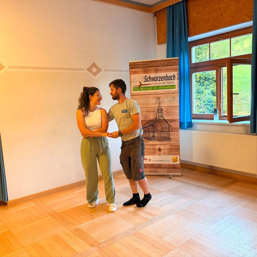 Ein Mann und eine Frau stehen in einem Raum mit Holzboden und sehen sich an. Der Mann trägt ein graues T-Shirt und Shorts, während die Frau ein weißes Tanktop und grüne Hosen trägt. Hinter ihnen steht ein Holzschild mit dem Text 'Schwarzenbach'.