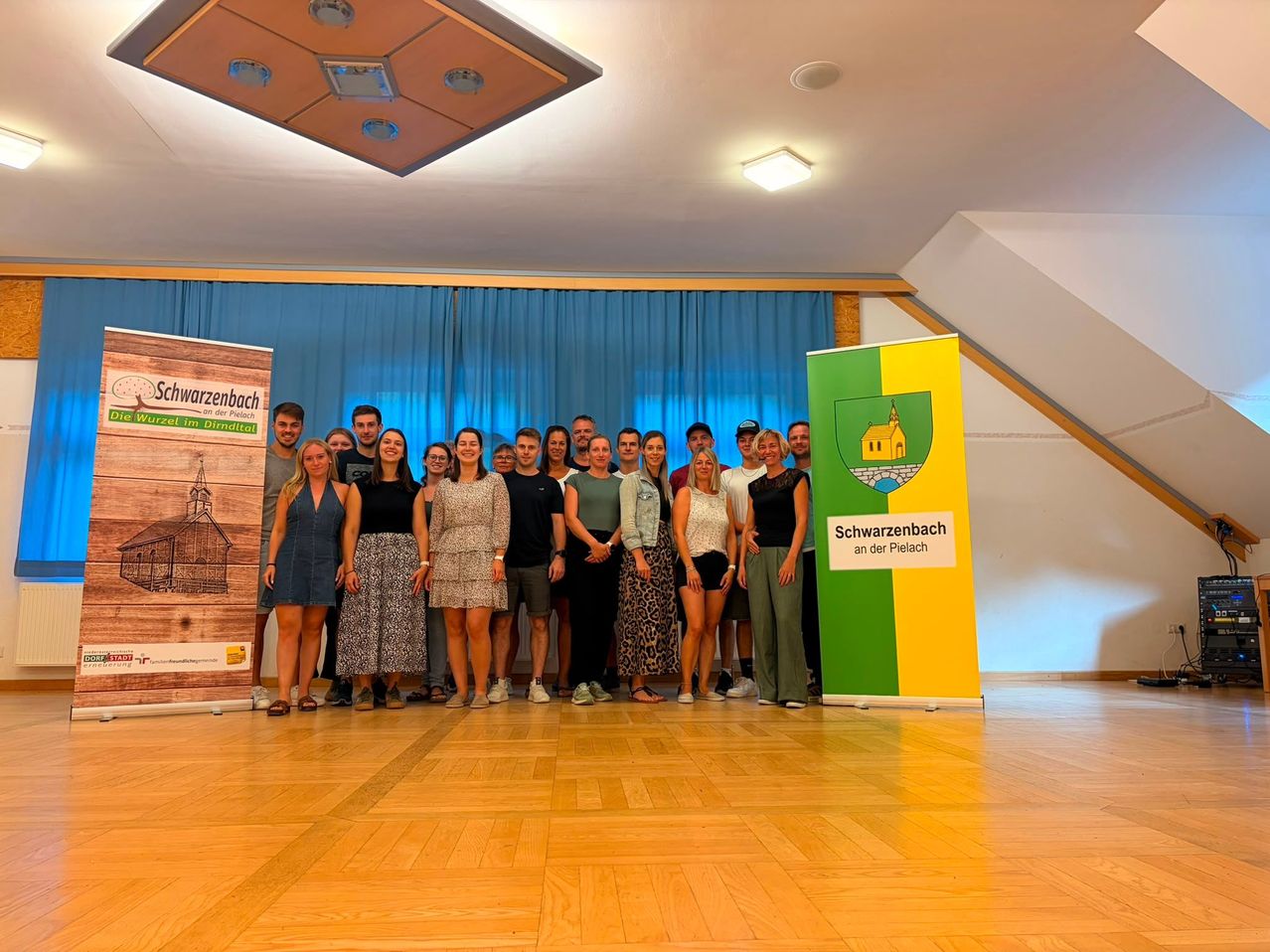 Eine Gruppe von Menschen posiert für ein Foto in einem Raum mit Holzböden, blauen Vorhängen und Bannern. Die Banner haben den Text 'Schwarzenbach' und ein Logo.