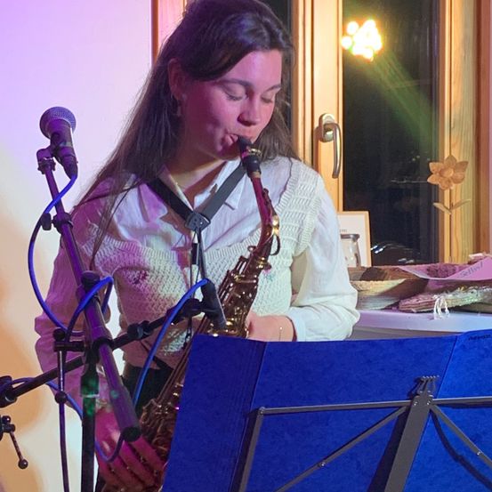 Eine Frau spielt die Saxophon vor einem Mikrofon. Sie steht in einem Raum mit einer Holztür und einer Blume am Fenster.