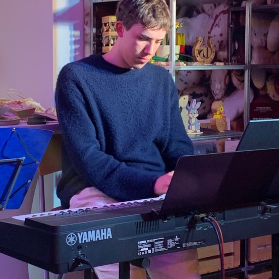 Ein Mann in einem blauen Pullover sitzt vor einem Yamaha-Keyboard, möglicherweise in einem Musikstudio, mit einem offenen Laptop vor ihm.