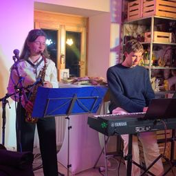 Eine Frau spielt Saxophon und ein Mann spielt Klavier, beide konzentriert auf ihre Instrumente schauend. Sie stehen vor einem Mikrofon und einem Notenständer. Dahinter befindet sich ein Raum mit Regalen voller Spielzeug und einem pinken Licht von einer Lampe.