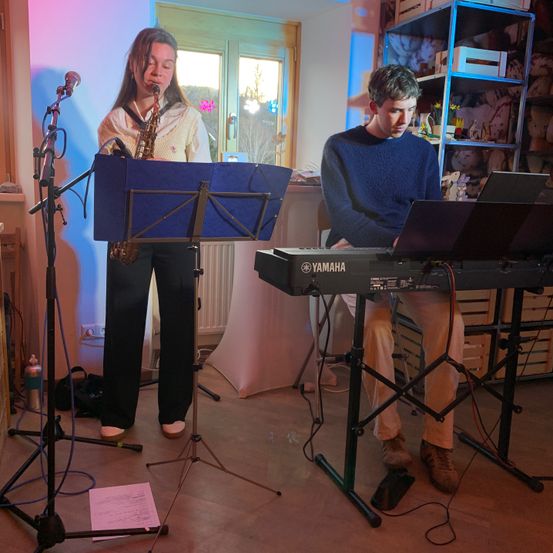 Zwei Personen spielen Musik in einem Raum mit lila und rosa Licht. Eine Frau spielt Saxophon, während ein Mann Klavier spielt.