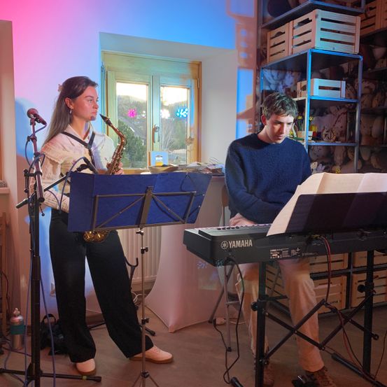Eine Frau in Pullover und Hose spielt Saxophon. Ein Mann sitzt auf einem Stuhl und spielt Klavier. Sie sind in einem Raum mit Fenster, Regalen und einer rosa Wand.
