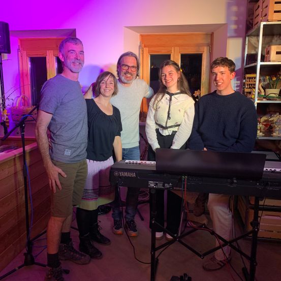 Eine Gruppe von fünf Personen steht in einem Raum mit lila Beleuchtung. Vier von ihnen lächeln und einer schaut zur Seite. Sie sind wahrscheinlich eine Band, und einer von ihnen spielt Klavier.