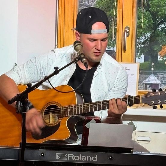Ein Mann spielt Gitarre und Keyboard in einem Raum mit einem Fenster, das auf Bäume und ein kleines Haus blickt. Er trägt eine schwarze Kappe und ein weißes Hemd.