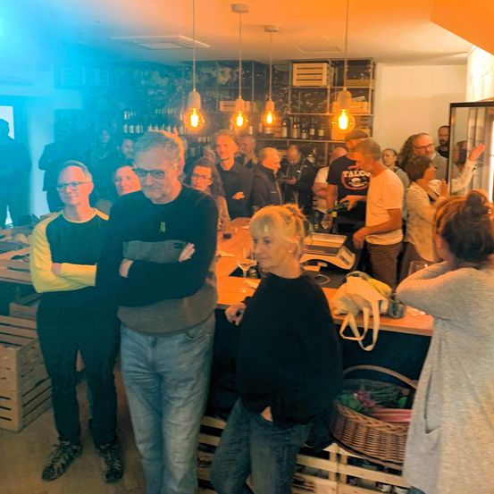 Eine Gruppe von Menschen, einschließlich Erwachsener, befindet sich in einer Bar. Einige stehen, andere sitzen. Eine Frau mit blondem Haar steht vor einer Theke. Flaschen und andere Gegenstände sind auf den Regalen.