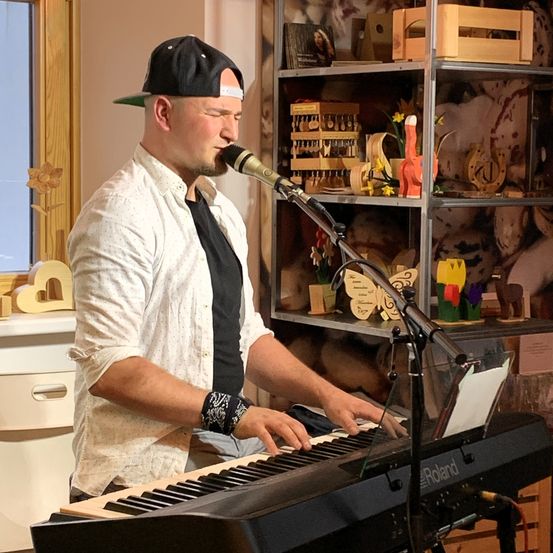 Ein Mann mit einer Kappe spielt eine Tastatur vor einem Mikrofon, mit Regalen voller Holzspielzeug im Hintergrund.