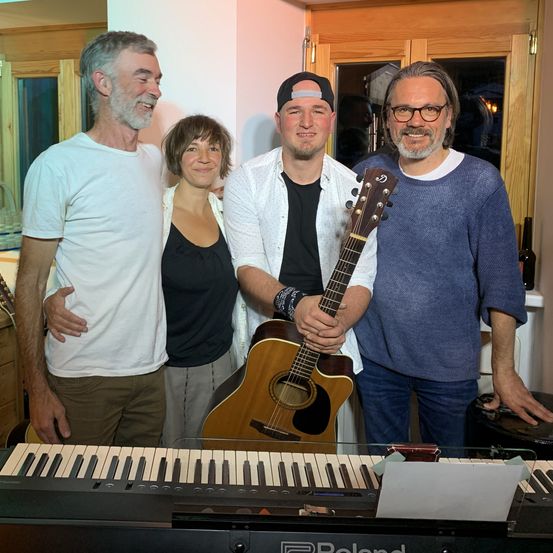 Vier Personen stehen in einem Raum mit Musikinstrumenten, lächeln und posieren für ein Foto. Ein Mann hält eine Gitarre, während ein anderer Mann neben einem Klavier steht. Die Frau und ein weiterer Mann stehen hinter ihnen.