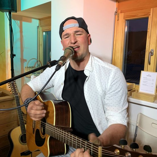 Ein Mann in einer schwarzen Kappe und einem weißen Hemd spielt eine Gitarre, während er in ein Mikrofon singt. Eine zweite Gitarre ruht auf einer Holzkiste zu seiner Linken. Hinter ihm ist ein offenes Fenster zu sehen, mit einem Schild auf der Fensterbank.