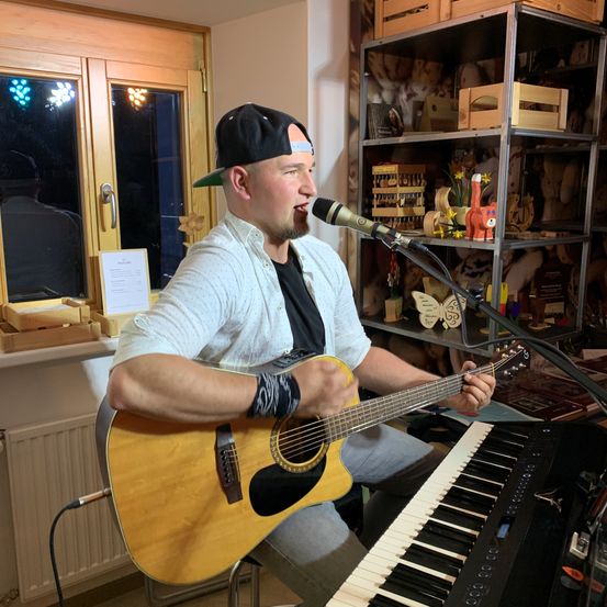 Ein Mann spielt Gitarre und singt in ein Mikrofon in einem Raum mit Fenster, Regalen und Spielzeug.