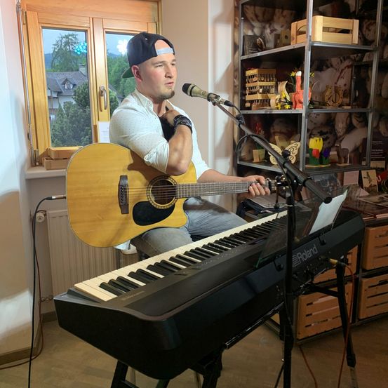 Ein Mann spielt Gitarre und Klavier und singt in ein Mikrofon in einem Raum mit einem Holzfenster und Regalen mit verschiedenen Gegenständen.