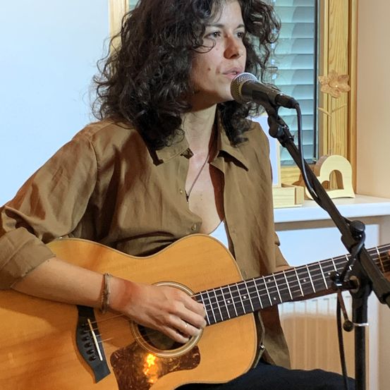 Eine Frau mit lockigem Haar spielt Gitarre und singt in ein Mikrofon. Sie trägt ein braunes Hemd und eine Halskette. Hinter ihr befindet sich ein Fenster mit Jalousien.