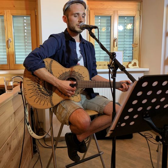 Ein Mann in einem blauen Hemd und Hut spielt eine Gitarre mit Sonnendesign, auf einem Hocker vor einem Mikrofon sitzend. Die Umgebung umfasst eine Holztheke, ein Glas Wasser und einen Notenständer mit einem Notenblatt.