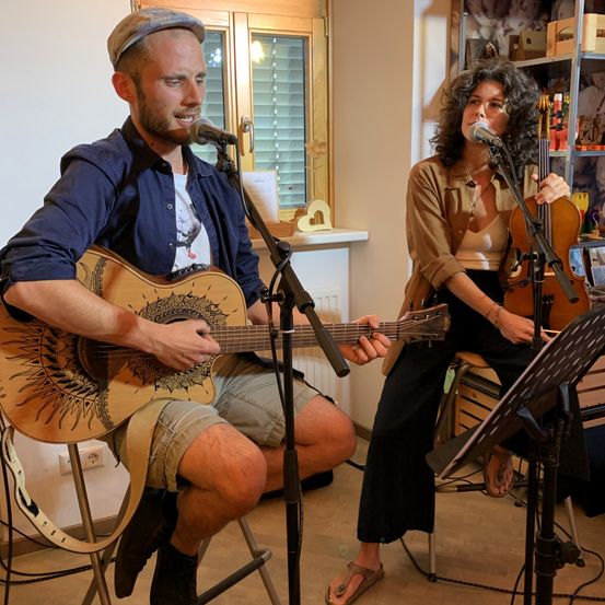 Ein Mann und eine Frau führen ein Lied auf. Der Mann spielt Gitarre, während die Frau Geige spielt. Beide singen in Mikrofone. Sie befinden sich in einem Raum mit einem hölzernen Fenster und Regalen.