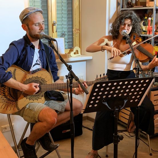 Ein Mann und eine Frau spielen Musik in einem Raum. Der Mann spielt Gitarre und singt in ein Mikrofon, während die Frau Geige spielt. Sie sitzen beide auf Stühlen.