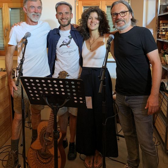 Vier Menschen stehen gemeinsam für ein Foto, zwei Männer und zwei Frauen, mit einer Gitarre und Mikrofonen davor. Sie scheinen Musiker zu sein.