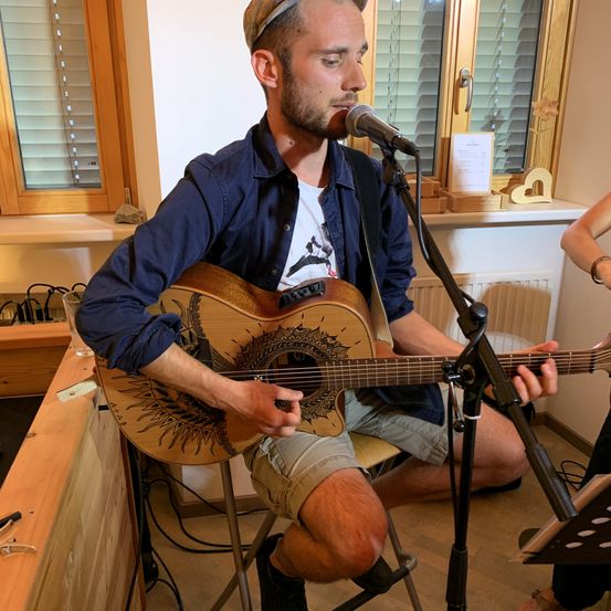 Ein Mann sitzt auf einem Stuhl und spielt eine Gitarre mit einem Mikrofon vor sich. Er trägt einen Hut und ein blaues Hemd. Hinter ihm befindet sich ein Fenster mit geschlossenen Jalousien.